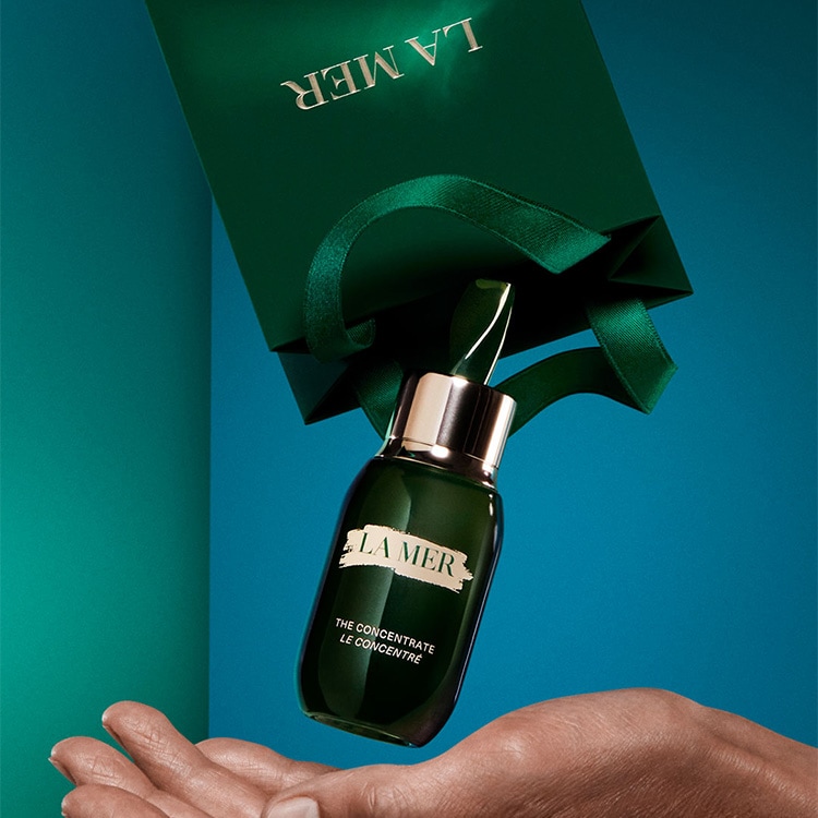 la mer product falling out of a green gift bag into a hand, on a blue green background